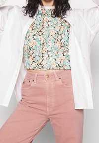 Chemise blanche boutonnée portée par-dessus un haut à motifs floraux verts, roses et bleus ; associée à un jean rose pastel avec des poches avant.