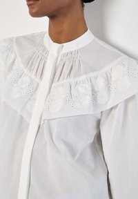 Weiße Baumwollbluse mit hohem Kragen, ausgestattet mit einer bestickten Passe mit floralen Mustern und Rüschendetails an den Schultern.