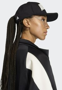 Gorra negra con logo blanco y correa ajustable, llevada con una chaqueta negra que presenta un panel blanco en contraste. Largos cabellos trenzados caen por detrás.