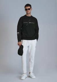 Zwarte sweatshirt met witte lijnen, gecombineerd met witte tapered broeken. Model houdt een zwarte pet vast en draagt witte sneakers. Neutrale achtergrond.