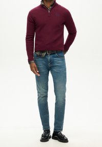 Mann, der einen burgunderfarbenen Reißverschluss-Pullover über einem karierten Hemd, mit blauen Jeans und schwarzen Quasten-Schuhen vor einem weißen Hintergrund steht.