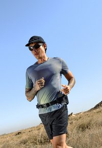 Läufer mit Sonnenbrille, Kappe und Tattoos joggt auf einem trockenen grasigen Weg unter klarem blauem Himmel.