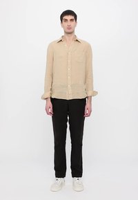 Camicia di lino beige chiaro a maniche lunghe con taschino sul petto, abbottonatura frontale e polsini arrotolati; abbinata a pantaloni neri e sneakers bianche.