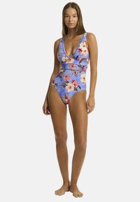 Model in een bloemen-eenstuk badpak met een diepe V-hals, staand op een witte achtergrond, die het ontwerp en de kleuren van de zwemkleding toont.