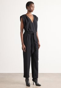 Mono negro con mangas cortas, escote en V, cintura ceñida y pantalones ajustados, complementado con botines de tacón y accesorios mínimos.