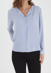 Blouse bleu clair à manches longues avec décolleté en V, en tissu lisse, offrant une coupe décontractée et un ourlet droit. Associée à un pantalon noir.
