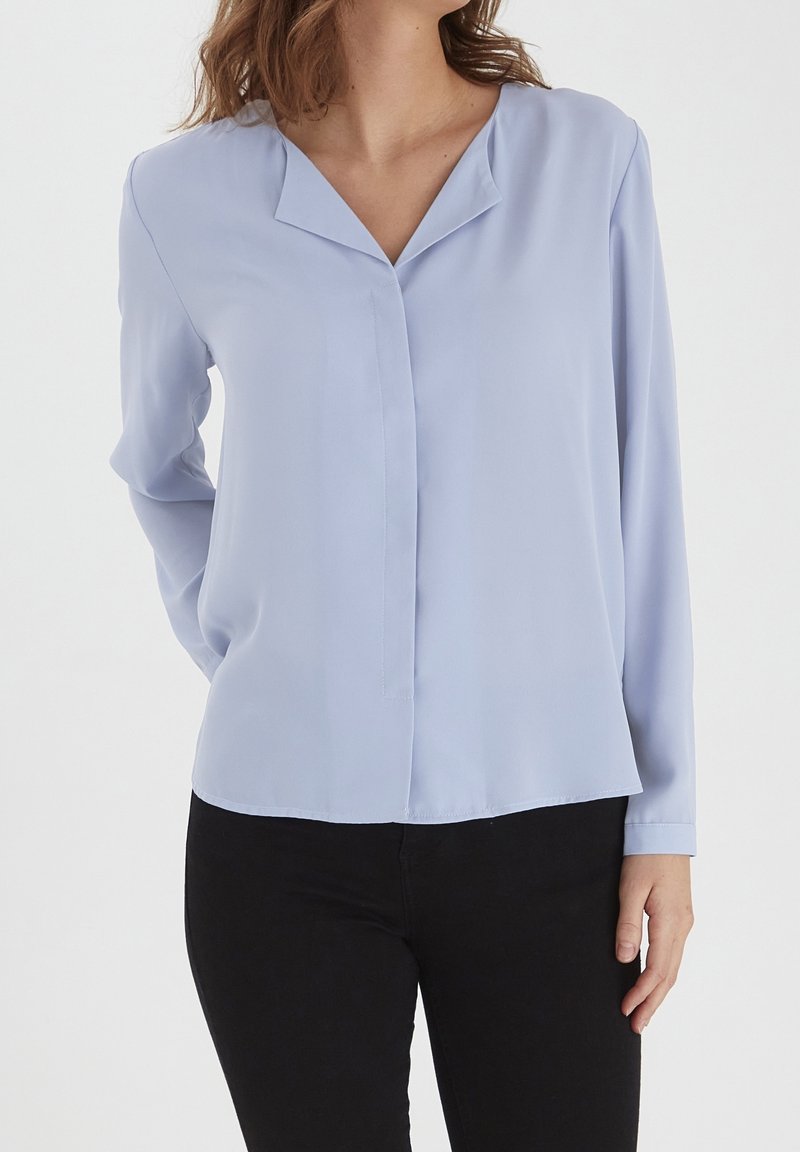 Blouse bleu clair à manches longues avec décolleté en V, en tissu lisse, offrant une coupe décontractée et un ourlet droit. Associée à un pantalon noir.