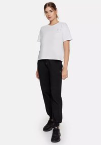 Witte t-shirt met korte mouwen en een relaxed fit, voorzien van een kleine grafische afbeelding op de borst, gecombineerd met zwarte sweatpants en zwarte sportschoenen.