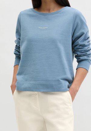 Personne portant un sweat-shirt bleu clair à manches longues avec le logo "Marc O'Polo" et un pantalon écru, les mains dans les poches.