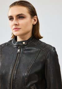 Giacca in pelle nera con chiusura a zip, colletto alto e spalle imbottite. Presenta una texture liscia e leggermente invecchiata.