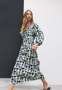 Robe à manches longues avec un motif géométrique bleu et crème, dotée d'une taille froncée, d'un décolleté en V et d'une jupe fluide. Tissu léger.