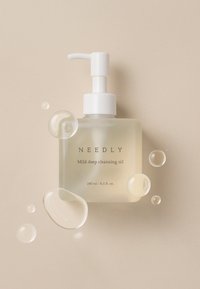 Przezroczysta szklana butelka z pompką, zawierająca olejek do oczyszczania oznaczony napisem „NEEDLY”, z białą pompką, jasnożółtym płynem i kilkoma przejrzystymi pęcherzykami.