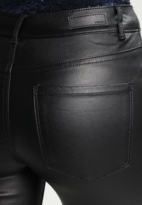 Mustan keinonahkaiset leggingsit, joissa on sileä pinta, takatasku ja ommeltu yksityiskohta vyötäröllä. Brändin tunniste näkyvissä.