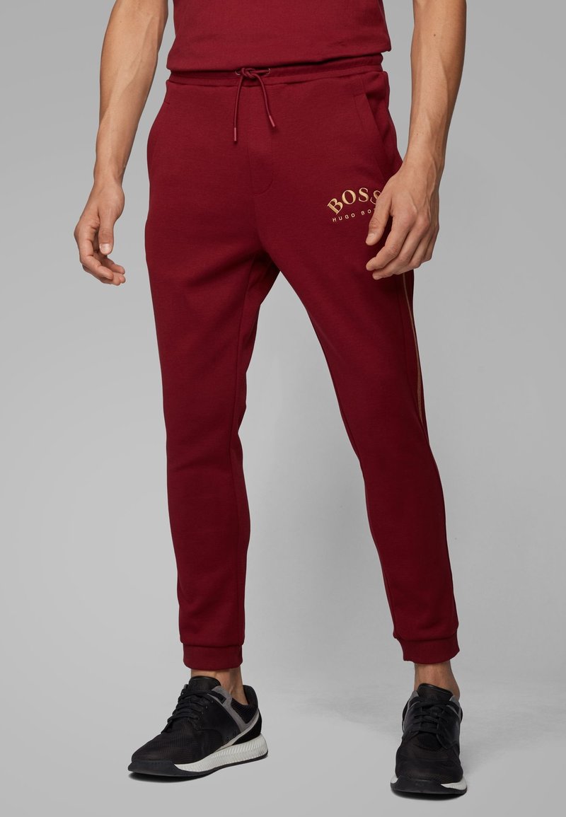 Pantalones deportivos burdeos con bolsillos laterales, cintura elástica y cordón; con texto de logo dorado. Fabricados con tela suave y texturizada.