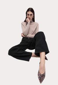 Béžový rolák, černé kalhoty s širokými nohavicemi a lodičky s leopardím potiskem. Modelka sedí se zkříženýma rukama, neutrální pozadí.