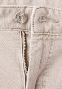 Pantalones de mezclilla beige con un zipper plateado, botón de presión metálico con grabado y tejido texturizado. Vista en primer plano de los cierres.