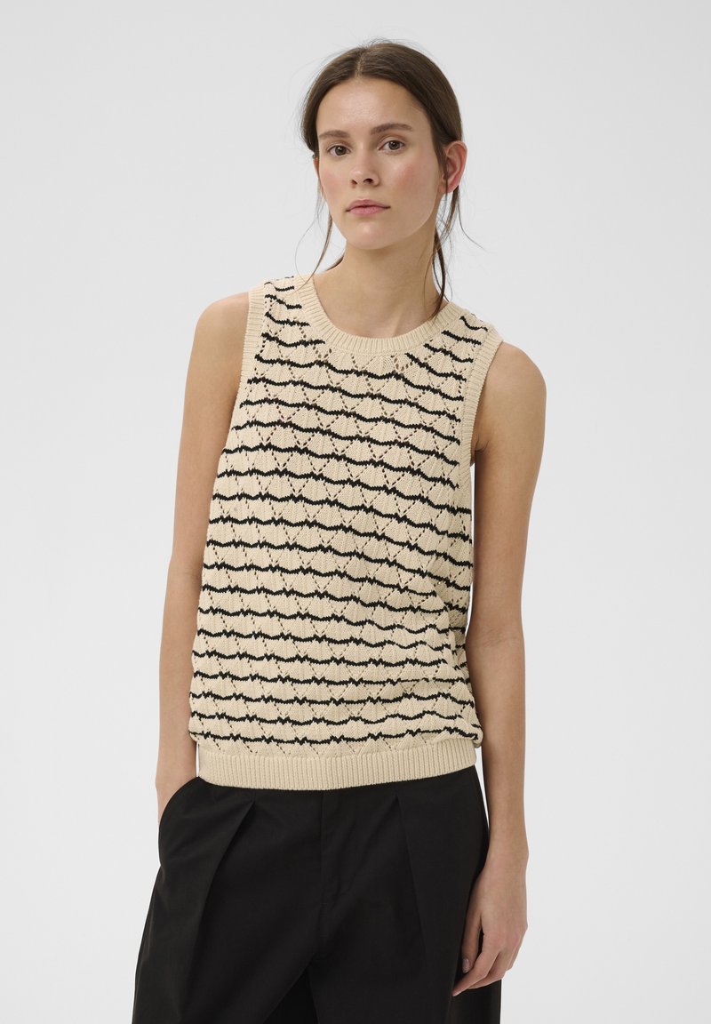 Jeune femme aux cheveux bruns portant un haut tricoté sans manches beige avec des motifs ondulés noirs et un pantalon noir, se tenant devant un fond uni.