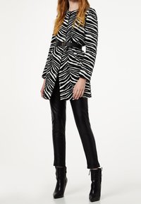 Mujer vestida con abrigo de estampado de cebra en blanco y negro con cinturón negro, pantalones de cuero negros y botines negros contra un fondo blanco.