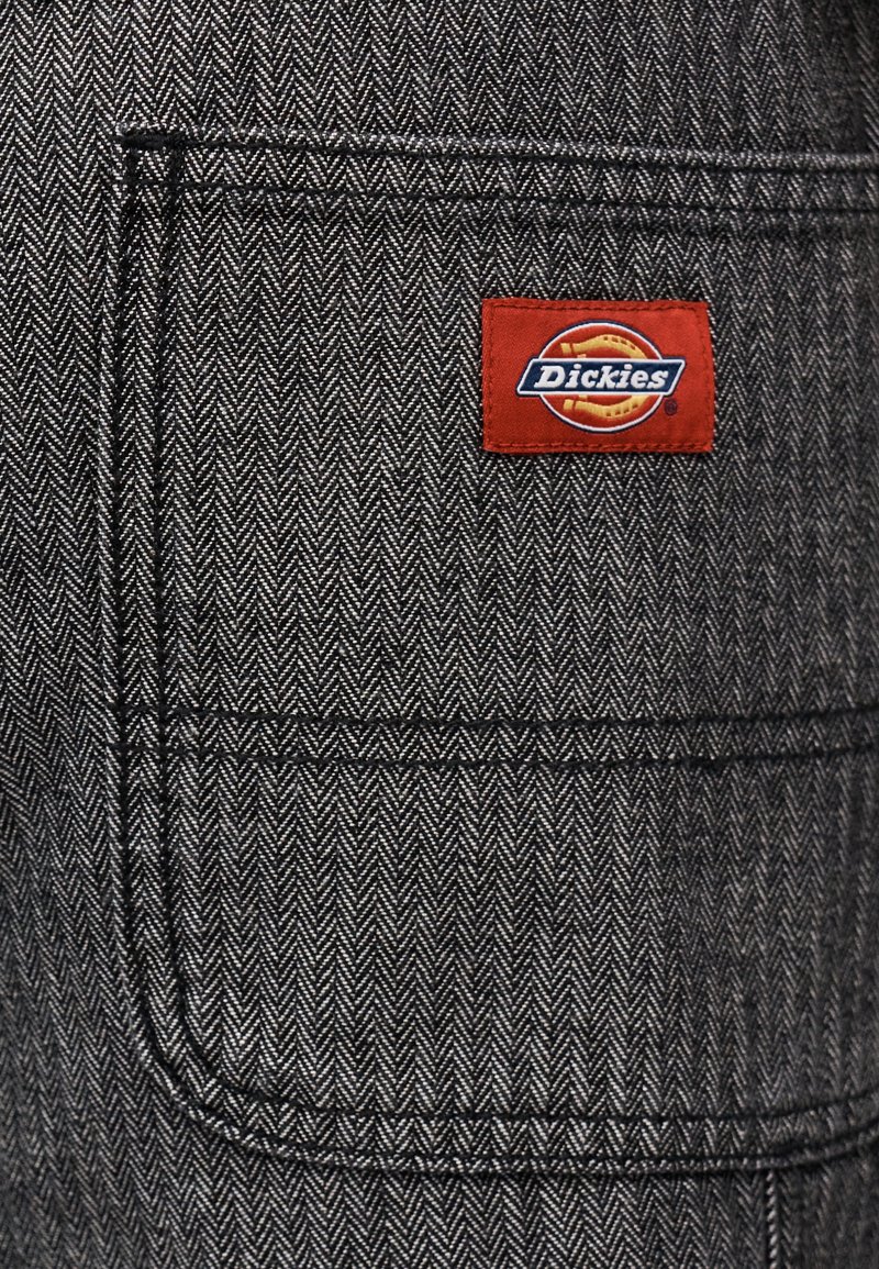 Primo piano di tessuto grigio a spina di pesce con una tasca cucita e un'etichetta rettangolare rossa con la scritta "Dickies" sull'area della tasca in alto a destra.