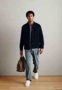 Crna jakna, siva košulja, svijetlo plave traperice i bijele tenisice; nosi smeđu kožnu torbu. Drveni pod i jednobojna pozadina.