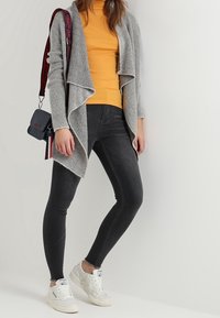 Grauer gestrickter Cardigan, gelber Rollkragenpullover, dunkle Jeans und weiße Sneaker. Als Accessoire eine kleine graue Umhängetasche mit gestreiftem Tragegurt.