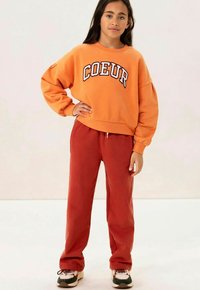 Oranje sweatshirt met "COEUR" in witte letters, gecombineerd met rode sweats. Tan en zwarte sneakers maken de casual outfit compleet.