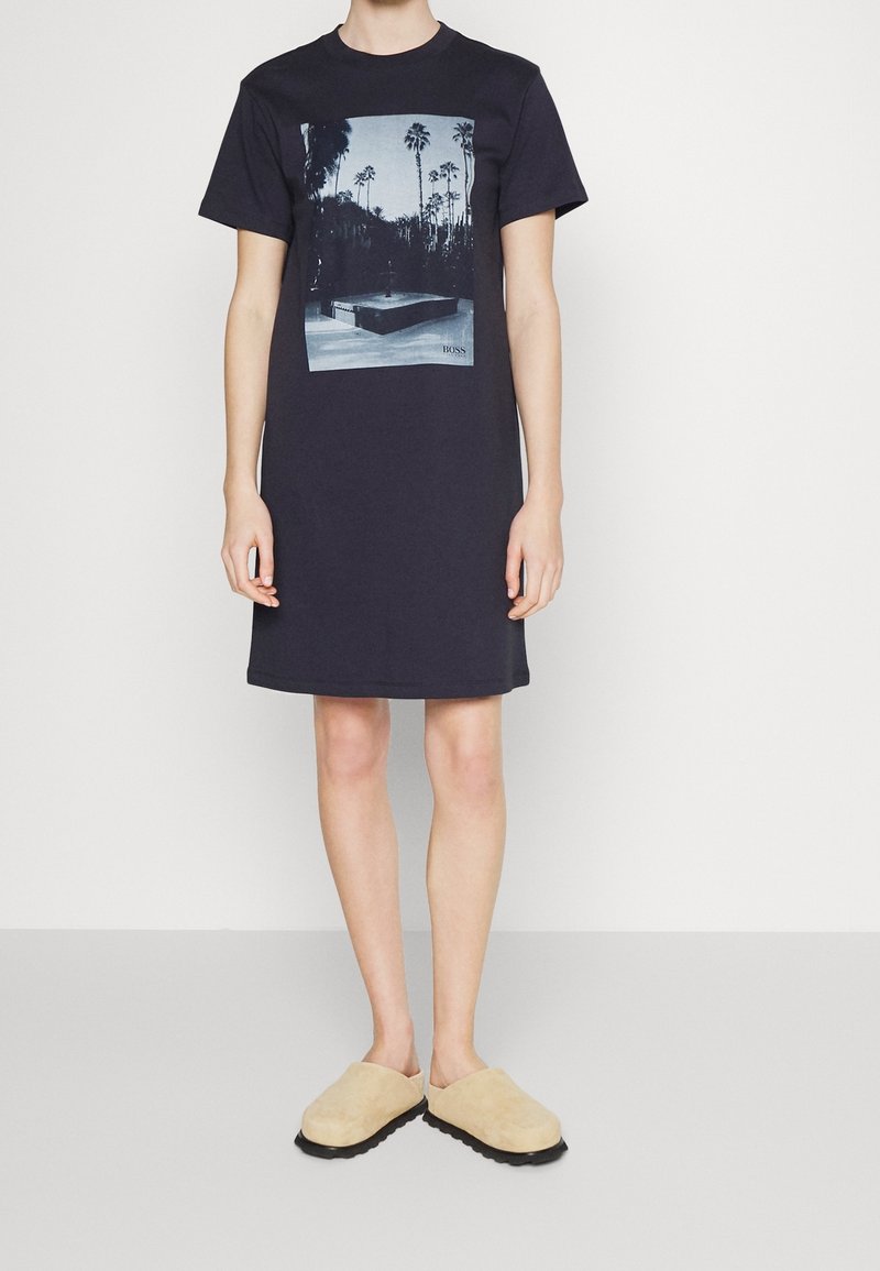 Personne portant une robe noire à manches courtes avec un imprimé palmiers noir et blanc et des chaussures beiges à enfiler, debout sur un fond blanc.