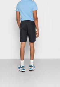 Tummat shortsit suoraa mallia, yhdistettynä vaaleansiniseen lyhythihamaiseen paita. Valkoiset sukat ja sinisiä yksityiskohtia sisältävät lenkkarit täydentävät asua.