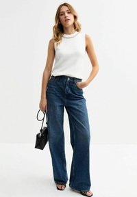 Vit ärmlös topp med dekorerad halsringning, kombinerad med höga vida blå denimjeans. En svart handväska och sandaler fulländar looken.