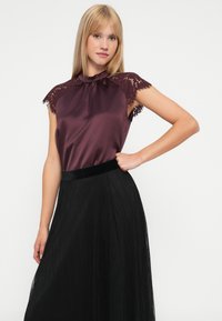 Camicia di satin borgogna con maniche a cappuccio in pizzo e scollatura arricciata, abbinata a una gonna nera plissettata con una vita in velluto.