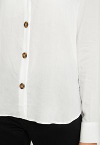 Camicia bianca in cotone con vestibilità comoda, caratterizzata da cinque bottoni marroni e maniche lunghe con polsini abbottonati. Tessuto dalla consistenza morbida.