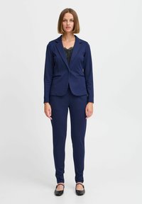 Námořnický oblek obsahující přizpůsobený blazer s klopami a slim-fit kalhoty, doplněný černým krajkovým topem pod ním a černými balerínami.