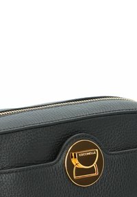 Zwarte leren tas met een gestructureerd oppervlak, voorzien van een opvallend goudkleurig rond logo en een ritssluiting aan de bovenkant.