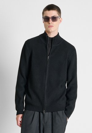 Mann mit dunklen Sonnenbrillen, einem schwarzen, gerippten Reißverschluss-Pullover und grauen plissee Hosen steht vor einem schlichten weißen Hintergrund.