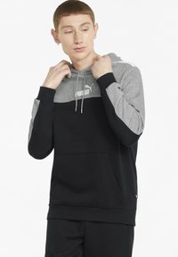 Sudadera con capucha que tiene una sección superior gris y una sección inferior negra. Presenta un bolsillo tipo canguro y el logo de PUMA en blanco en el pecho.
