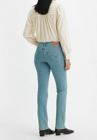 Jean en denim bleu clair avec une taille haute, poches arrière comportant un logo distinctif, associé à un blouse crème et des bottes noires.