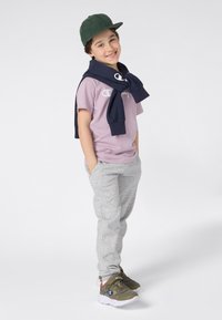 Niño con una camiseta de color púrpura claro con un logo, pantalones de chándal grises y zapatillas verdes oliva, con una sudadera azul marina atada alrededor del cuello.