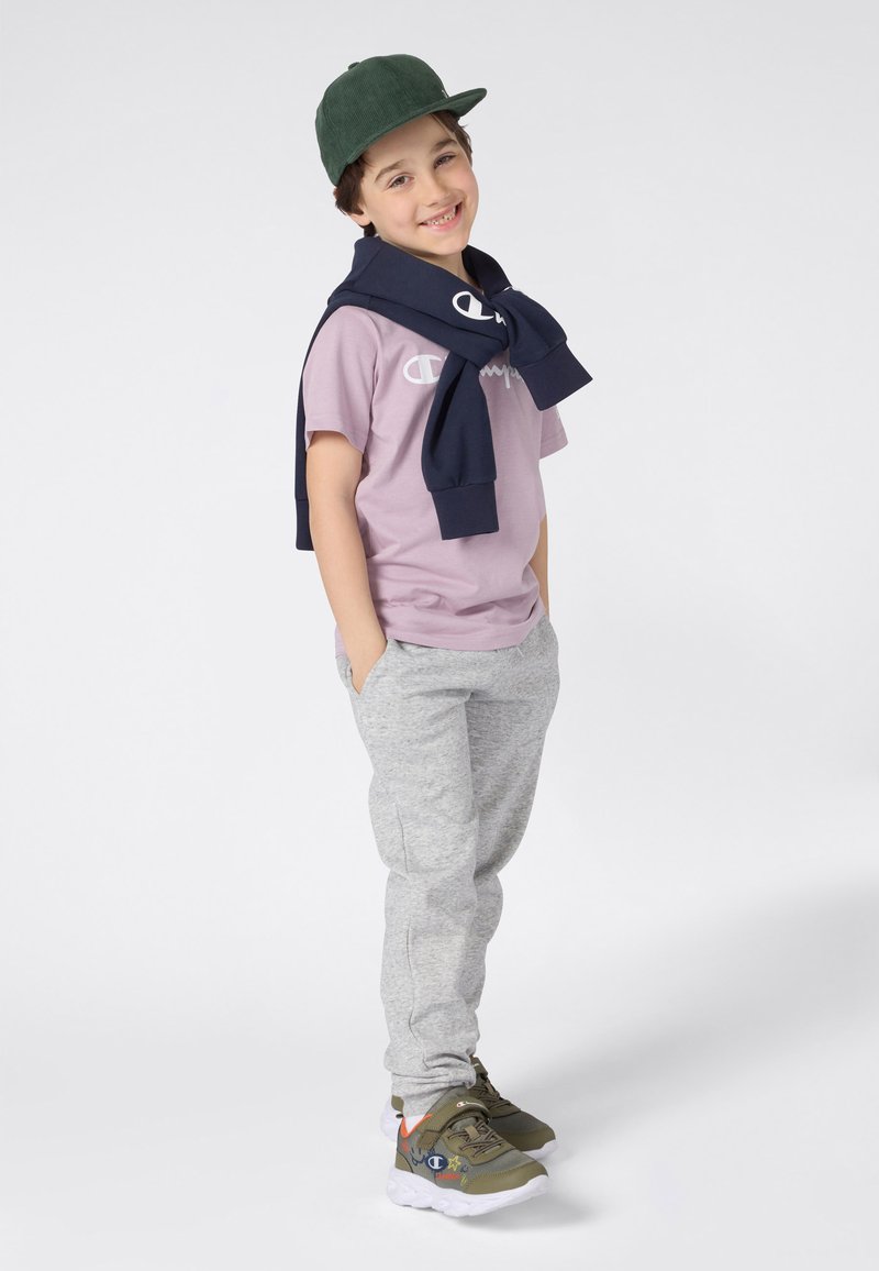 Niño con una camiseta de color púrpura claro con un logo, pantalones de chándal grises y zapatillas verdes oliva, con una sudadera azul marina atada alrededor del cuello.