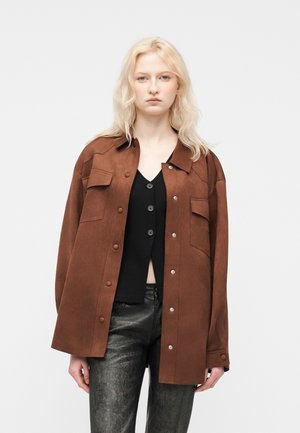 Blondynka w dużej brązowej kurtce z guzikami na czarnej bluzce zapinanej na guziki oraz ciemnych, teksturowanych spodniach, stojąca na jednolitym jasnym tle.