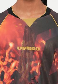 Artimiausias planas žmogaus, dėvinčio multicolorinį sportinį marškinį "Umbro", su oranžinės, juodos ir geltonos abstrakčios ugnies raštu.