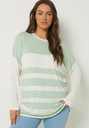 Kobieta o długich brązowych włosach nosi luźny miętowo-biały sweter w paski oraz jasnoniebieskie dżinsy, stoi na jednolitym tle.