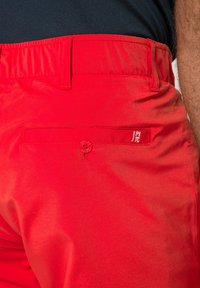Rode shorts met een gladde textuur hebben een elastische tailleband, een achterzak met knoopsluiting en een klein logo aan de zijkant.