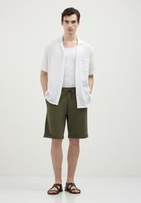 Giovane uomo con capelli ricci scuri che indossa una camicia bianca aperta a maniche corte, una canotta bianca, pantaloni corti verde oliva e sandali marroni scuri.