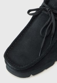 Black suede ankle boot with a rounded toe, light stitching details, and black shoelaces. The sole is textured with a slight grip pattern.