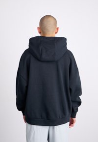 Oversized black hooded sweatshirt with textured fabric, dropped shoulders, ribbed hem, and back yoke detail. Paired with light gray pants.