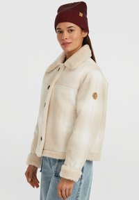 Beige ternetjakke med en blød, faux fur krave og manchetter. Brune knapper og et rundt logo patch på ærmet. Iført med en vinrød hue.