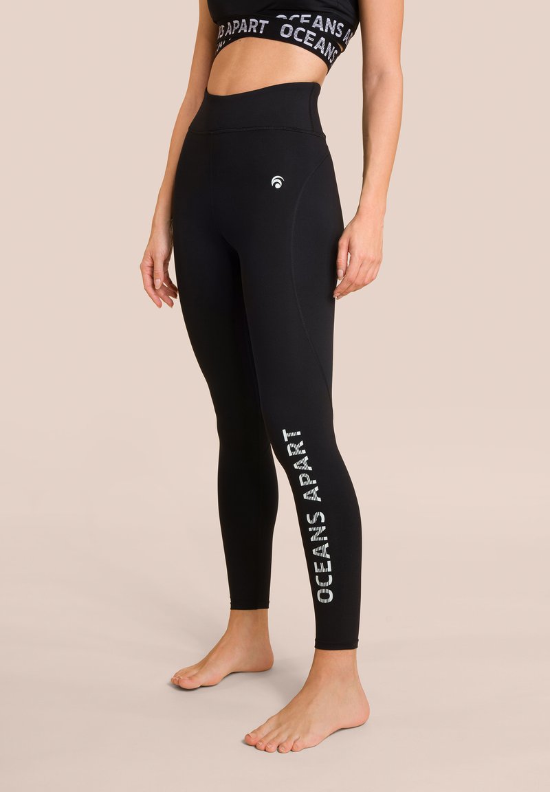 Leggings neri a vita alta realizzati in materiale elasticizzato, con la scritta "OCEANS APART" stampata in bianco lungo la gamba sinistra e un logo sulla cintura.