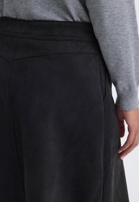 Person trägt einen schwarzen Wildlederrock und einen grauen Langarmpullover, zeigt die Seitennaht und das Taschen-Detail des Rocks.