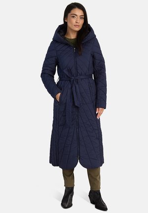 Naine, kellel on seljas pikk tumesinine kapuutsiga tikitud mantel vöököitega, oliivisääre püksid ja mustad pahkluukingad, seisab valgel taustal.