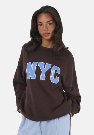 Femme aux longs cheveux foncés portant un sweat-shirt marron foncé avec de grandes lettres bleues "NYC" et un pantalon bleu clair sur un fond uni.